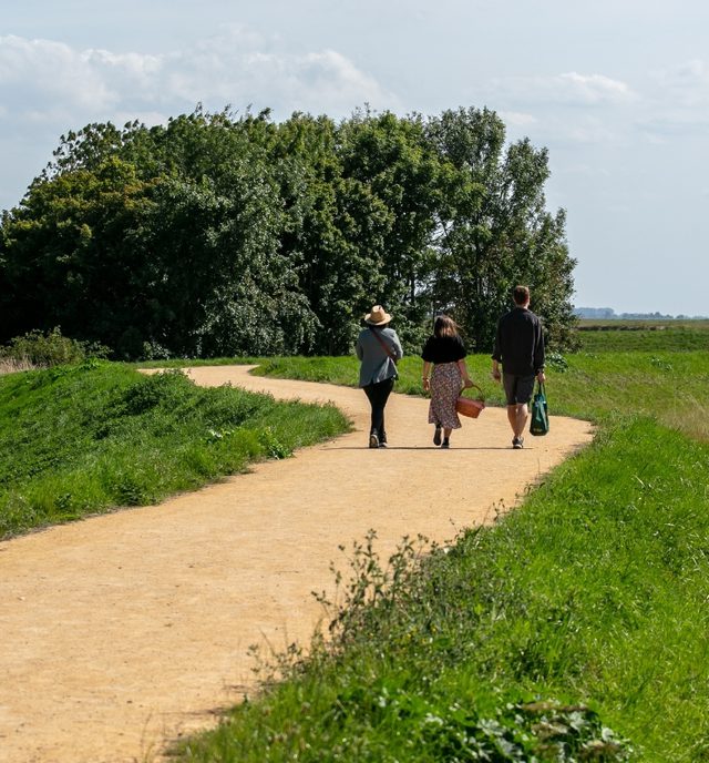 All weather path with 3 people walking along the middle, with a picnic basket in hand under clear blue skies surrounded by grass.